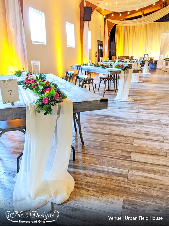 Bright and summery garland of tropical and wild flowers in deep purples and oranges run with spiked wild greenery atop a flowing Cream chiffon runner, on top of a Long Wood table.